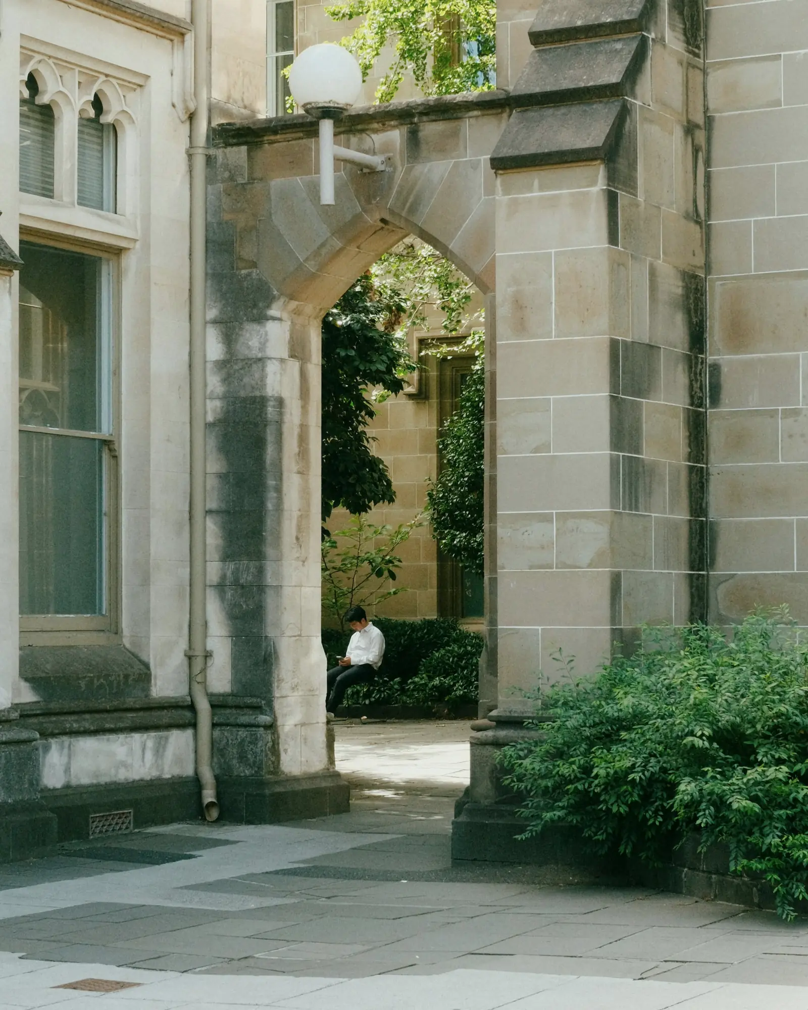 墨尔本大学的石砌拱门下，一名学生坐在庭院的绿植旁看手机